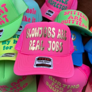 May include: A collection of neon-colored trucker hats with various phrases in gold lettering. The hats are pink, green, blue, and yellow. Some phrases include "BLOWJOBS ARE REAL JOBS" and "HURTING FOR A SQUIRTING".