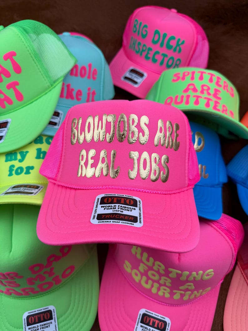 May include: A collection of neon-colored trucker hats with various phrases in gold lettering. The hats are pink, green, blue, and yellow. Some phrases include "BLOWJOBS ARE REAL JOBS" and "HURTING FOR A SQUIRTING".