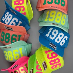 May include: A collection of colorful trucker hats with the year "1986" printed on the front. The hats come in various colors, including pink, blue, green, and yellow, with contrasting text colors. The hats have a mesh back and a curved brim.