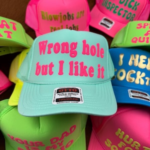 May include: A collection of trucker hats in various bright colors, including pink, green, and blue. Each hat features bold text in contrasting colors, with phrases like "Wrong hole but I like it" and "Big Dick Inspector". The hats have a mesh back and a snap closure.