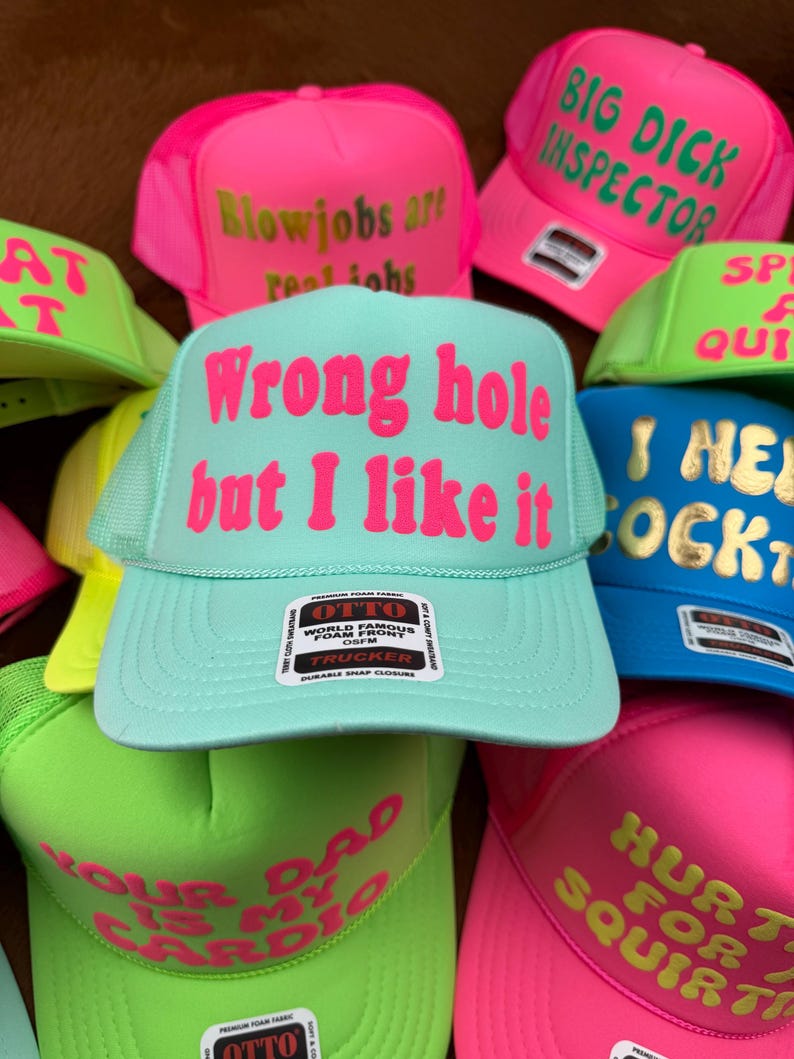 May include: A collection of trucker hats in various bright colors, including pink, green, and blue. Each hat features bold text in contrasting colors, with phrases like "Wrong hole but I like it" and "Big Dick Inspector". The hats have a mesh back and a snap closure.