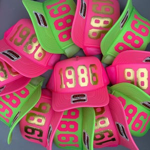 以下が含まれることがあります： ネオンピンクとネオングリーンのトラッカーキャップのコレクション。各キャップのフロントフォームパネルには、メタリックゴールドのフォントで「1986」とプリントされています。キャップは構造化されたフロント、メッシュのバック、つばに小さな長方形のラベルが付いています。
