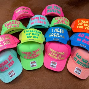 Könnte beinhalten: Eine Sammlung von Trucker-Caps in verschiedenen Farben, darunter Pink, Grün und Blau. Jede Kappe zeigt einen kühnen, humorvollen Text in kontrastierenden Farben. Die Caps haben einen Mesh-Rücken und einen gebogenen Schirm.