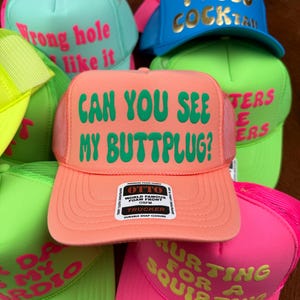 May include: A collection of trucker hats in various neon colors, including pink, green, and blue. Each hat features bold text in contrasting colors, with phrases like "Can You See My Buttplug?" and "I Need Cocktails."