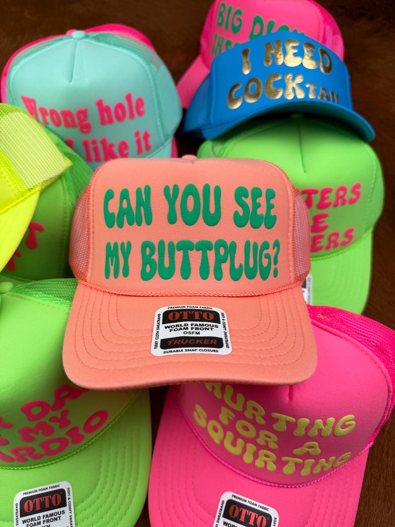 May include: A collection of trucker hats in various neon colors, including pink, green, and blue. Each hat features bold text in contrasting colors, with phrases like "Can You See My Buttplug?" and "I Need Cocktails."