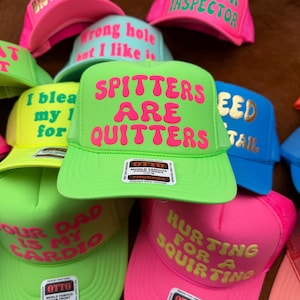 May include: A collection of trucker hats in various neon colors, including pink, green, and blue. Each hat features bold, humorous text in contrasting colors. The hats have a mesh back and a foam front.