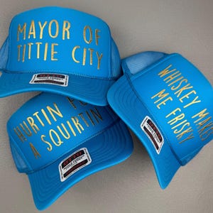 May include: Three bright blue trucker hats with mesh backs and gold lettering. The hats feature the text MAYOR OF TITTIE CITY, WHISKEY MAKES ME FRISKY, and HURTIN FOR A SQUIRTIN. Each hat has a small rectangular label on the brim with Otto branding.