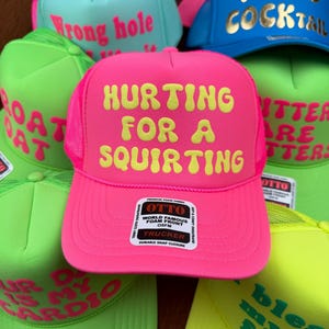 May include: A collection of trucker hats in various neon colors, including pink, green, and blue. Each hat features bold, humorous text in a contrasting color. The hats have a mesh back and a snap closure.