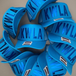 May include: A collection of bright blue trucker hats with the letters "KWLA" printed across the front. The hats have a mesh back and a curved brim. The hats are arranged in a circular pattern.