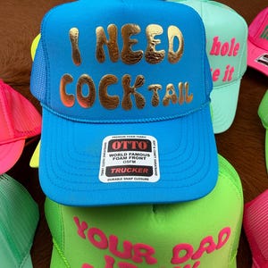 May include: A collection of trucker hats in various colors. The primary hat is bright blue with "I NEED COCKTAIL" in gold lettering. Other hats include neon green and pink, each with unique phrases.