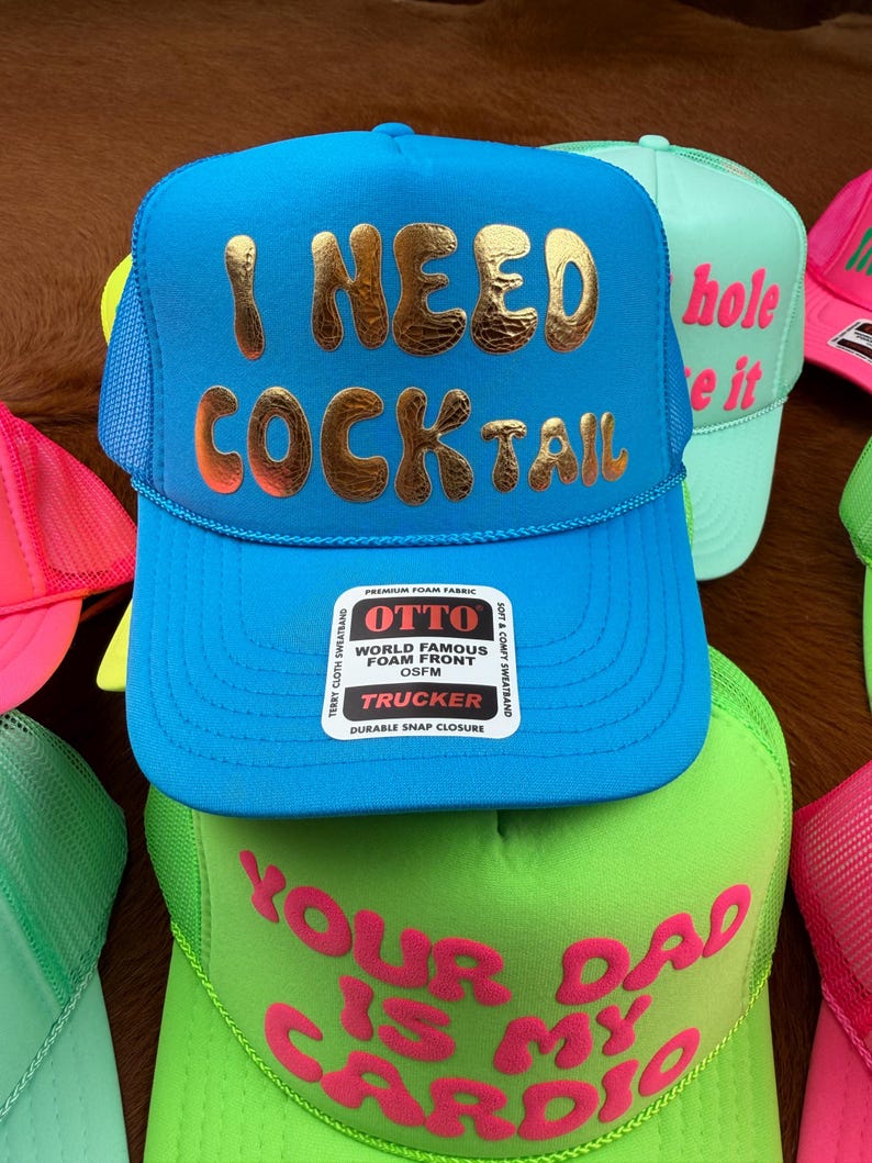 May include: A collection of trucker hats in various colors. The primary hat is bright blue with "I NEED COCKTAIL" in gold lettering. Other hats include neon green and pink, each with unique phrases.