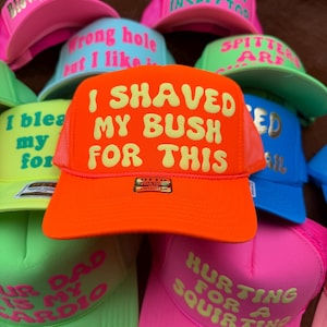 May include: A collection of colorful trucker hats with various phrases printed on the front. The hats are in shades of pink, green, blue, and orange. The text includes phrases like "I shaved my bush for this" and "Hurting for a squirting."