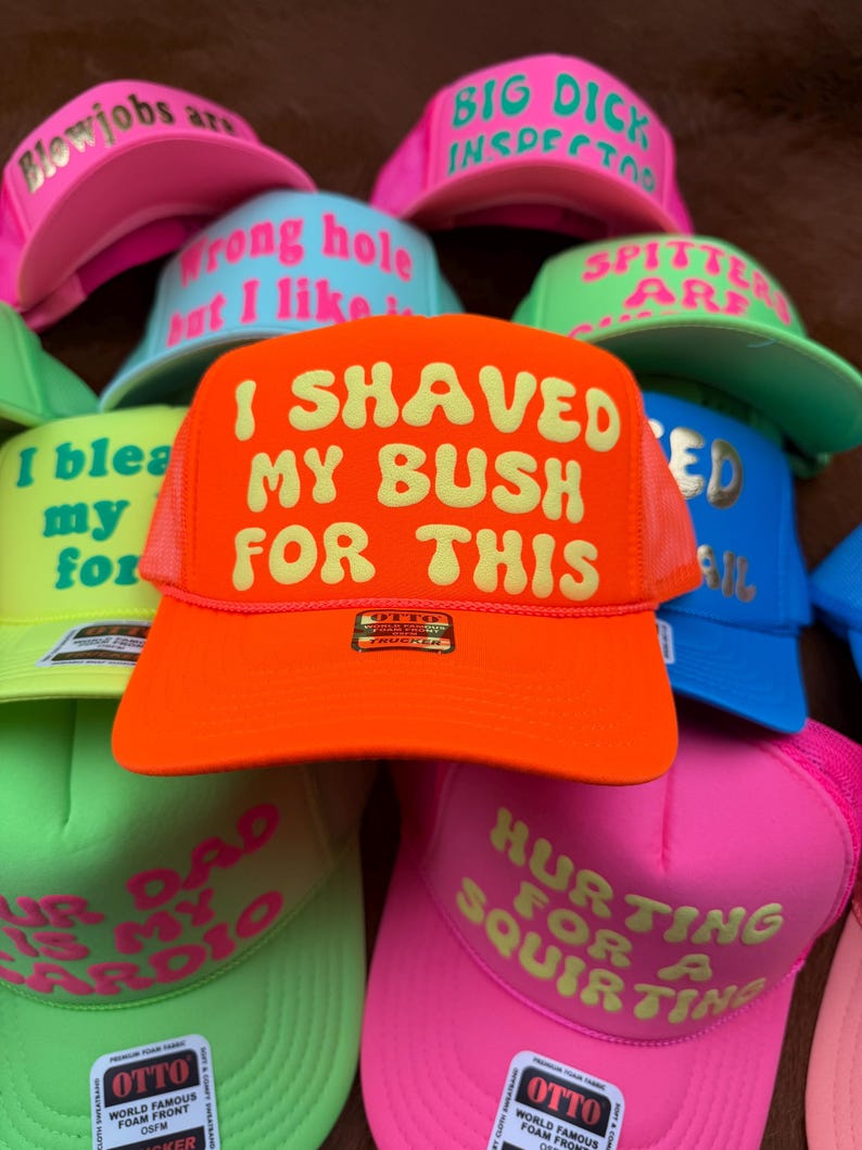 May include: A collection of colorful trucker hats with various phrases printed on the front. The hats are in shades of pink, green, blue, and orange. The text includes phrases like "I shaved my bush for this" and "Hurting for a squirting."
