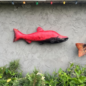 Puede incluir: Una vibrante escultura de pez de madera roja con detalles en negro, colgada contra una pared gris texturizada. El pez está suspendido por un hilo fino, mostrando sus aletas y cuerpo detallados. Luces decorativas están colgadas arriba.