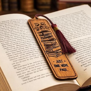 May include: Wooden bookmark with a tassel, resting on an open book. The bookmark features a stack of books and the phrase "JUST ONE MORE PAGE." The tassel is burgundy, and the bookmark is brown.