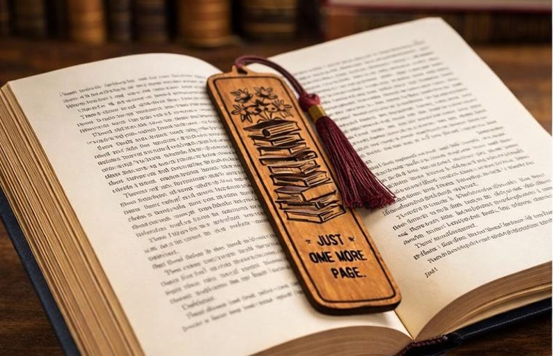 May include: Wooden bookmark with a tassel, resting on an open book. The bookmark features a stack of books and the phrase "JUST ONE MORE PAGE." The tassel is burgundy, and the bookmark is brown.