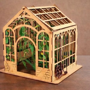 Op de afbeelding: Een miniatuur houten kas met ingewikkelde lasergesneden details. De structuur heeft gewelfde ramen, een latten dak en een kleine decoratieve haspel. Er is groen te zien aan de binnenkant, wat wijst op een tuinthema. Het algehele ontwerp is lichtbruin.