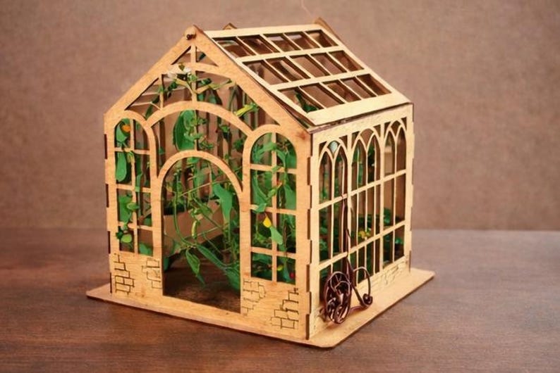 Op de afbeelding: Een miniatuur houten kas met ingewikkelde lasergesneden details. De structuur heeft gewelfde ramen, een latten dak en een kleine decoratieve haspel. Er is groen te zien aan de binnenkant, wat wijst op een tuinthema. Het algehele ontwerp is lichtbruin.