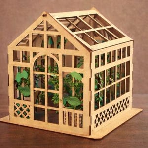 Op de afbeelding: Een miniatuur houten kas met een puntdak en transparante panelen. De structuur heeft ingewikkelde uitgesneden patronen en bevat kleine groene planten. De kas staat op een rechthoekige basis, met een natuurlijke houtkleur.