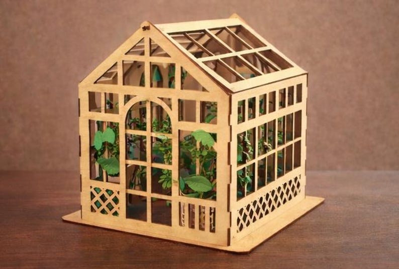 Op de afbeelding: Een miniatuur houten kas met een puntdak en transparante panelen. De structuur heeft ingewikkelde uitgesneden patronen en bevat kleine groene planten. De kas staat op een rechthoekige basis, met een natuurlijke houtkleur.