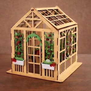 Op de afbeelding: Een gedetailleerde miniatuurhouten kas. De structuur heeft een zadeldak, ruiten en decoratieve bloemaccenten. Kleine witte plantenbakken met rode bloemen zijn aan de voorkant bevestigd. Het ontwerp is een charmante weergave van een tuinconstructie, gemaakt van hout.