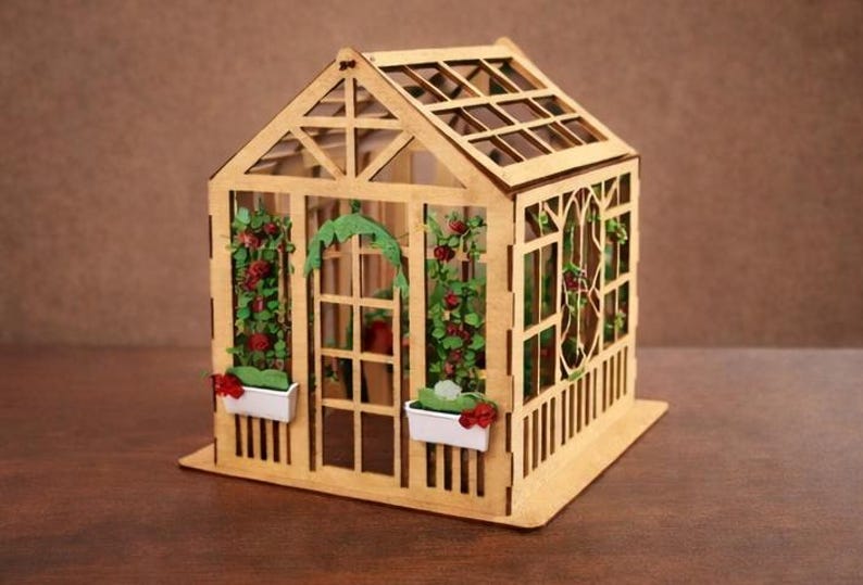 Op de afbeelding: Een gedetailleerde miniatuurhouten kas. De structuur heeft een zadeldak, ruiten en decoratieve bloemaccenten. Kleine witte plantenbakken met rode bloemen zijn aan de voorkant bevestigd. Het ontwerp is een charmante weergave van een tuinconstructie, gemaakt van hout.