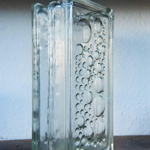 May include: A rectangular clear glass block featuring a textured surface with a pattern of raised circular bubbles of varying sizes. The block has a thick, transparent construction with smooth edges and a distinct grid-like design on the front face.