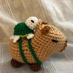May include: A tan crocheted capybara with dark brown feet and ears, featuring a small white and green crocheted turtle resting on its back, secured by two green yarn straps that wrap around the body of the capybara in a harness style.