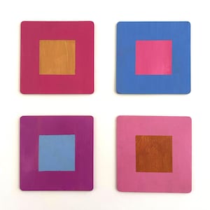 May include: Four square coasters with a square design. Each coaster has a different color combination: pink with gold, blue with pink, purple with blue, and pink with gold. The coasters are arranged in a square formation.
