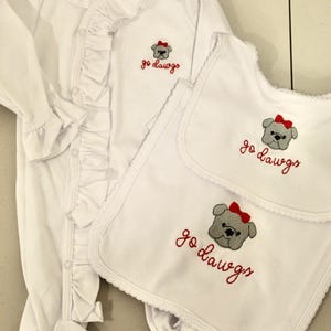 May include: A white long-sleeved footed sleeper with a ruffled front placket and two matching white bibs. Each item features an embroidered gray bulldog wearing a red bow with the red text "go dawgs" underneath. The sleeper has a Levi + Lane label.