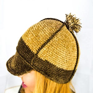 Puede incluir: Una gorra de estilo boina tejida a ganchillo con paneles alternos de hilo color canela y marrón oscuro. La gorra tiene una visera estructurada y un pequeño pompón texturizado en la corona. El diseño presenta un patrón de puntos visible.