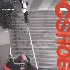 May include: A black, grey, and bright red athletic sneaker with a chunky sole and lace-up closure. The background features a black and white photograph of a skateboarder on a handrail, the text ROBDYRDEK, and large vertical orange text reading DCSHOECO.