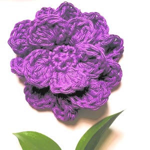 May include: A handmade, purple crochet flower with multiple layers of petals. The flower is made of thick yarn and has a textured appearance. A green leaf is visible in the foreground, set against a white background.