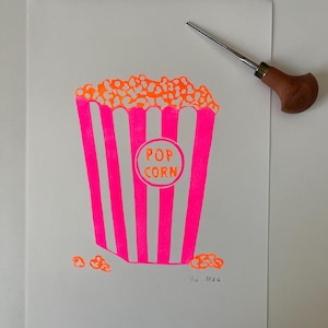 Peut inclure: Estampe linograveur rose et orange représentant un seau de pop-corn avec l'inscription "POP CORN" dans un cercle. Le pop-corn est orange, et le seau a des rayures roses. Un outil de gravure est posé sur le papier blanc.