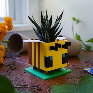 May include: A yellow and brown cubic bee-shaped planter with black eyes and white wings, featuring a green square base. A spiky green succulent plant is potted inside the hollow center of the bee figure, which has a blocky, pixelated design.