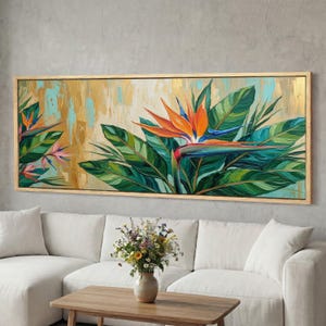 May include: A framed painting of a Bird of Paradise flower with orange, blue, and green hues. The artwork features a gold and teal background with lush green leaves. The painting is displayed above a white sofa and wooden coffee table.