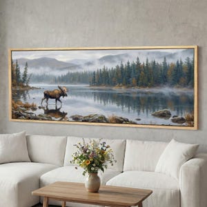 Peut inclure: Une impression murale panoramique représentant un élan brun pataugeant dans un lac calme et brumeux. La scène comprend une forêt dense de conifères le long du rivage, des montagnes en arrière-plan et des rochers dispersés dans l'eau.