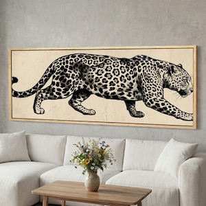 May include: A framed print depicting a black and white illustration of a leopard. The leopard, with its distinctive spotted coat, is set against a beige backdrop. The frame is light-coloured wood. The artwork is displayed above a white sofa.