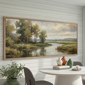 May include: A landscape painting in a light wood frame depicts a serene river scene. The artwork features lush green trees, grassy banks, and a cloudy sky, creating a tranquil atmosphere. The painting is displayed above a white table with decorative vases.