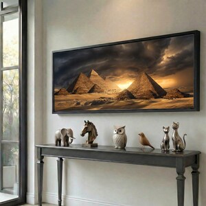 Op de afbeelding: Een ingelijste canvas print toont de piramides van Gizeh onder een dramatische lucht. Een donkergrijze console tafel toont decoratieve beeldjes: een olifant, een paardenkop, een uil, een vogel en twee kat sculpturen. De kamer heeft een neutraal kleurenpalet.