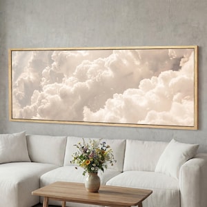 May include: A rectangular canvas print depicting a photograph of voluminous, white clouds with gentle, beige hues. The artwork is framed in a light-coloured wooden frame and hangs above a white sofa and wooden coffee table.