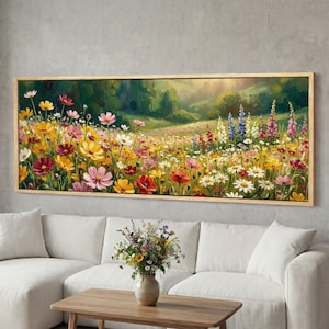 Op de afbeelding: Een ingelijst bloemenschilderij toont een levendige weide vol kleurrijke wilde bloemen. Het kunstwerk toont een verscheidenheid aan bloemen in tinten geel, roze, rood en wit, tegen een groene en zonnige achtergrond. Het schilderij is ingelijst in een licht houten frame.