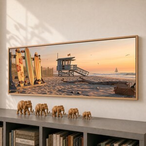 Op de afbeelding: Een ingelijste panoramische kunstdruk van een strandscène met drie surfplanken tegen een houten rek, een strandwachttoren, een zeilboot aan de horizon en een picknickmand. Daaronder staan vijf kleine bruine olifantenbeeldjes op een rij.