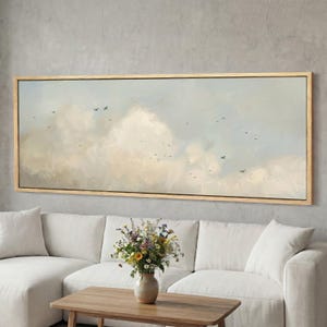 Op de afbeelding: Een groot rechthoekig ingelijst canvas kunstwerk dat een bewolkte lucht met vogels in vlucht afbeeldt. Het kunstwerk heeft zachte pastelkleuren van beige, blauw en wit. Het schilderij wordt tentoongesteld boven een witte bank en een houten salontafel.
