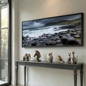 Puede incluir: Un cuadro paisajístico enmarcado de una costa tormentosa cuelga sobre una consola gris oscura. La mesa exhibe figuras decorativas, incluyendo un elefante, una cabeza de caballo, un búho, un pájaro y dos esculturas de gatos.