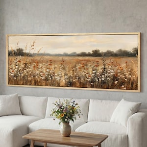 May include: A framed landscape painting depicts a field of wildflowers in shades of brown, orange, and white. The artwork is set against a neutral sky and framed in a light wood. The painting is displayed above a white sofa.