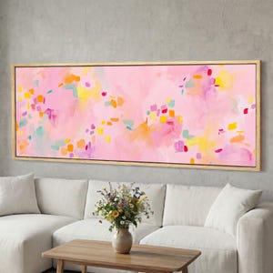 May include: Abstract art canvas with a pink background and colorful brushstrokes in yellow, orange, red, and teal. The artwork is framed in a natural wood frame and hangs above a white sofa and wooden coffee table.