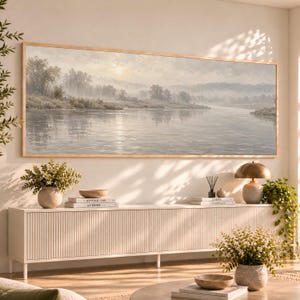 May include: A wide, rectangular landscape painting in a light wooden frame depicting a misty river scene with trees along the banks in muted tones of beige, gray, and soft white, mounted on a wall above a long, white, fluted sideboard.