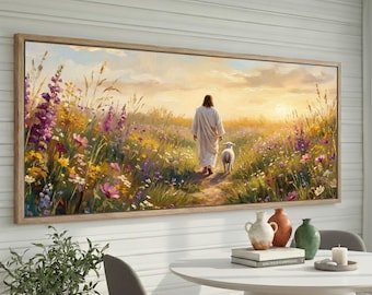 May include: A framed canvas print depicts a figure in a white robe walking with a lamb through a field of wildflowers. The artwork features a warm color palette with a sunset in the background. The frame is made of light-colored wood.