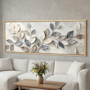 Può includere: Un'opera d'arte murale rettangolare con un rilievo di foglie stilizzate nei toni del bianco, grigio e marrone. L'opera è incorniciata in legno chiaro e appesa sopra un divano bianco. Il tema è la natura e l'arredamento della casa.